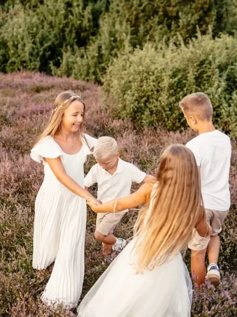 Vier Kinder in weißen Kleidern fassen sich in der blühenden Heide an den Händen.